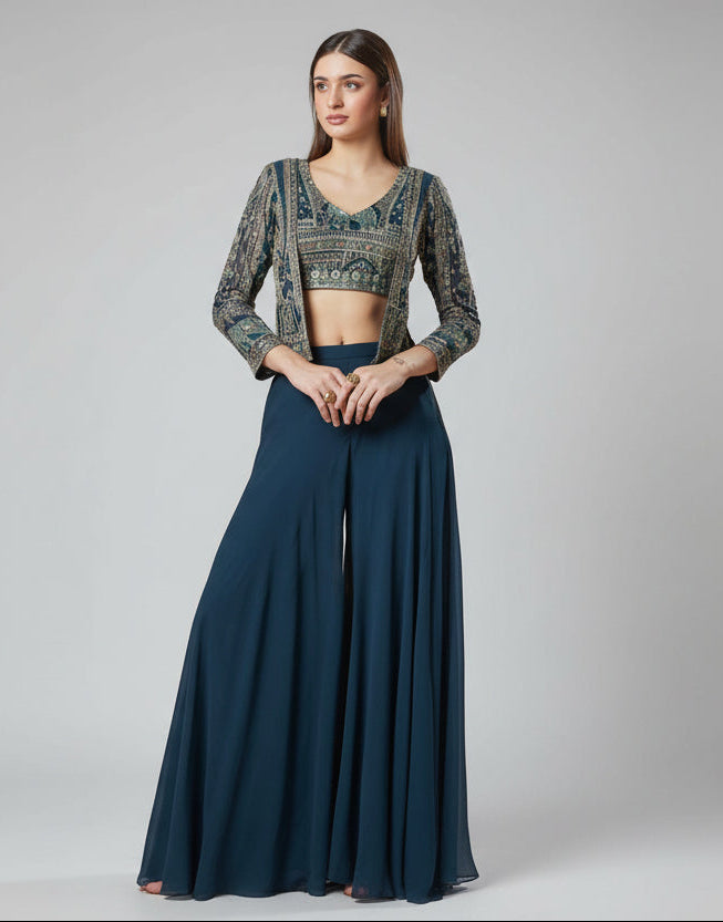 Woman wearing a blue outfit with a floral patterned top and wide-leg pants, standing against a neutral background with flowers.